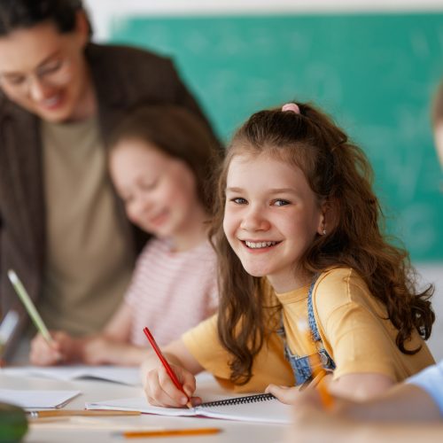 Smiling schoolgirl writing in a notebook while sitting at a classroom desk, with other children and a teacher blurred in the background.
