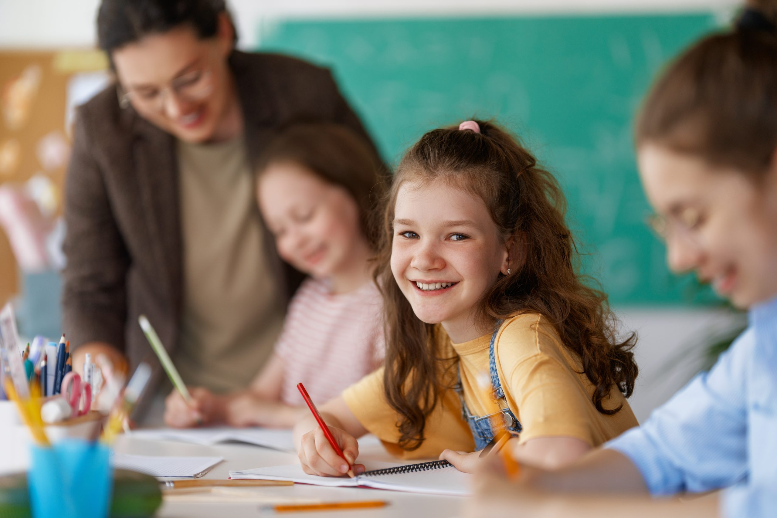 Smiling schoolgirl writing in a notebook while sitting at a classroom desk, with other children and a teacher blurred in the background.