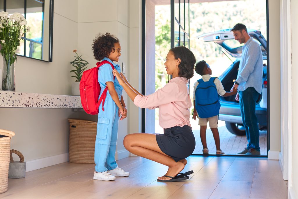 Mother helping her child with a red backpack get ready for school at home, while father and sibling wait by the car in the background.