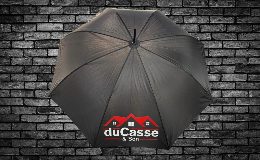 Black umbrella with the “duCasse & Son” logo printed in red and white, featuring a house roof design, displayed against a dark brick wall background.